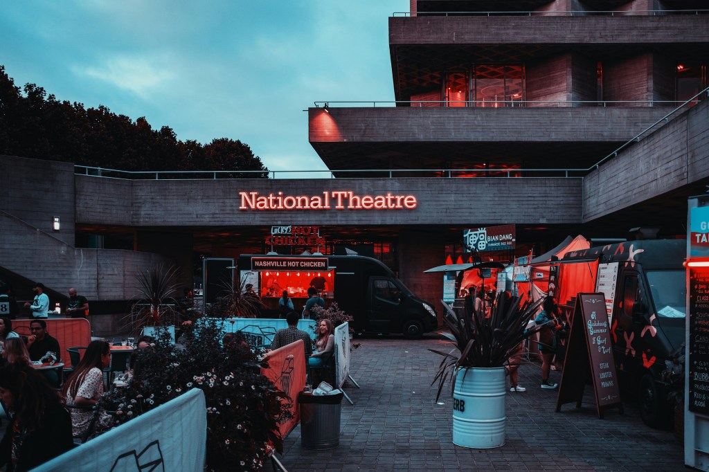 Exterior view of the National Theatre in London, featuring illuminated signage and food stalls in the foreground, with guests seated outdoors.